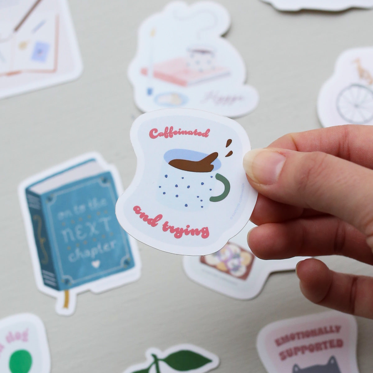 Hand holding a sticker with a coffee cup design and text, surrounded by other stickers on a light background.