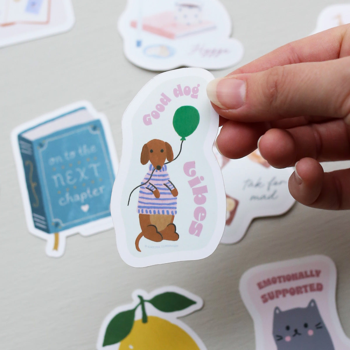 Hand holding a vinyl sticker with a dog, balloon, and the text 'good dog vibes', surrounded by other colorful stickers on a light surface.