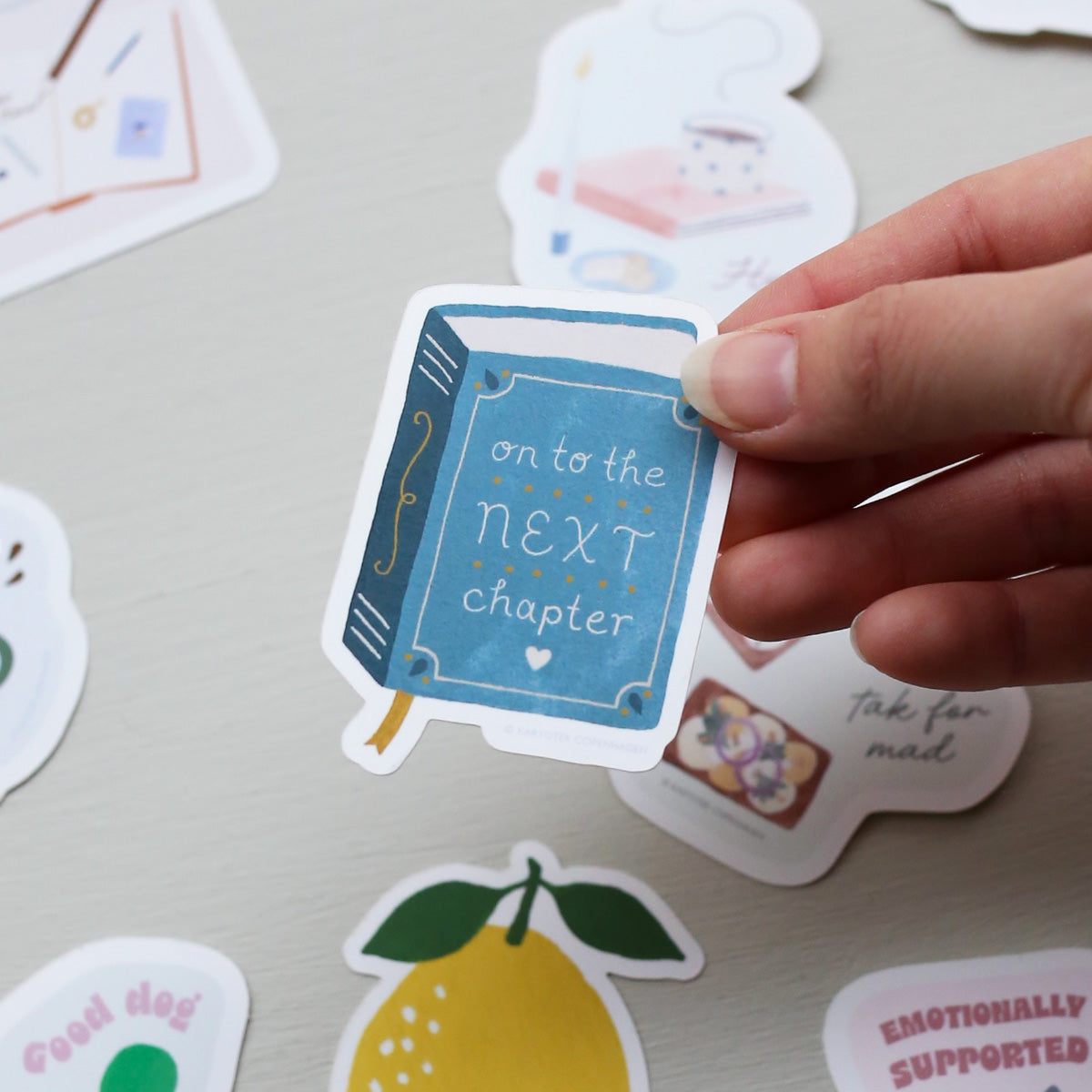 Hand holding a vinyl book-shaped sticker with 'On to the next chapter' text among other decorative stickers on a light surface.