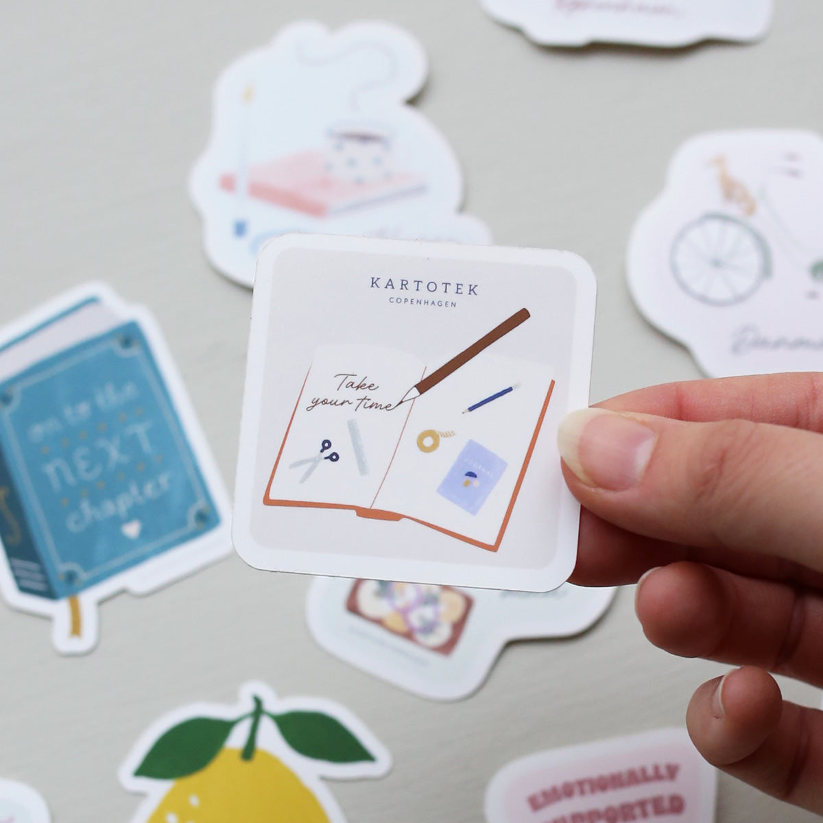 Hand holding a vinyl sticker with a journal filled with journaling illustrations, a pencil writing 'take your time' and a Kartotek Copenhagen logo, on a surface with other stickers.