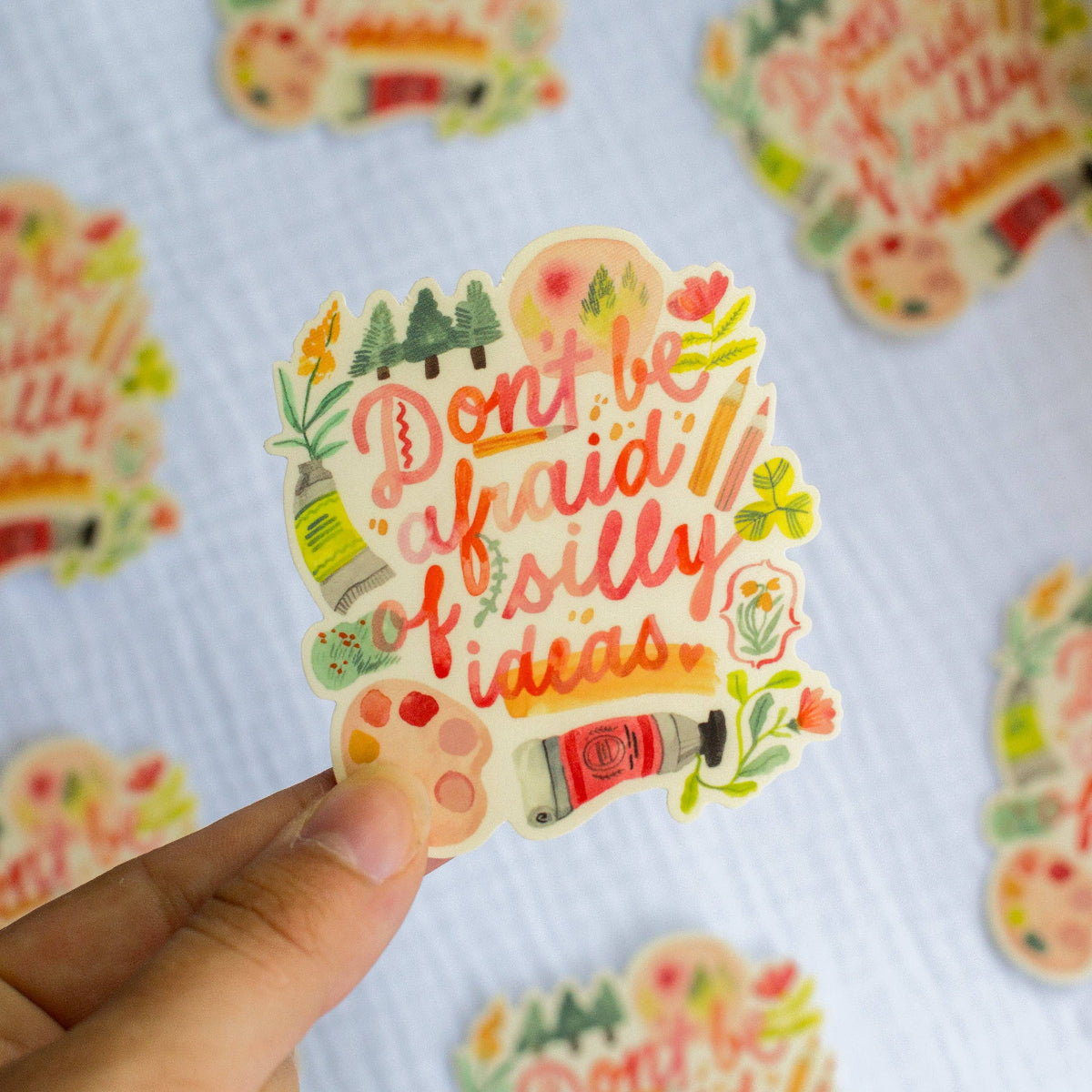 Colorful sticker with 'Don't be afraid of silly ideas' text held by a hand on a patterned background.