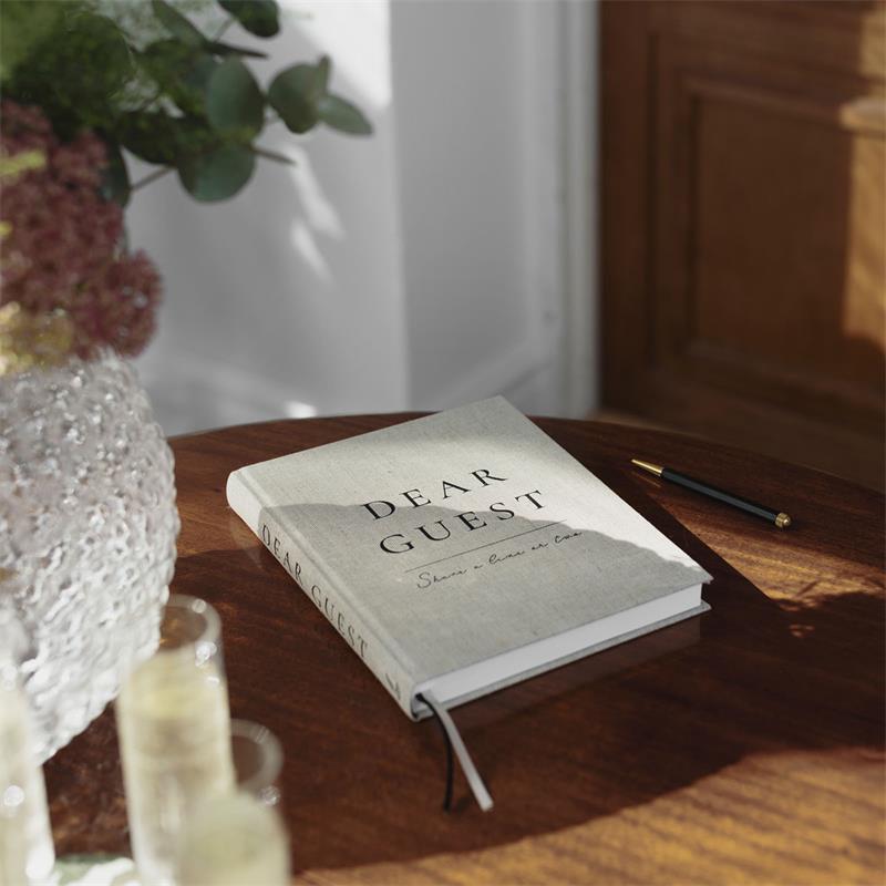 A hardcover journal titled 'DEAR GUEST' resting on a wooden table in a sunlit room, with a glass vase, flowers, and a pen nearby.