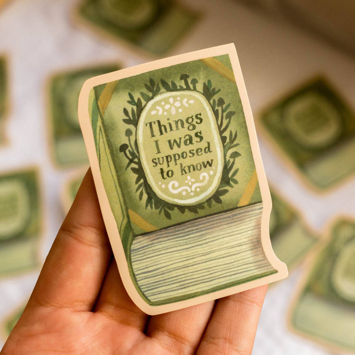 Hand holding a green book-shaped sticker with text 'Things I was supposed to know' against a blurred background of similar cards.