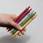 A hand holding five Kozo brand retractable pens in 5 different colors against a plain light gray background.