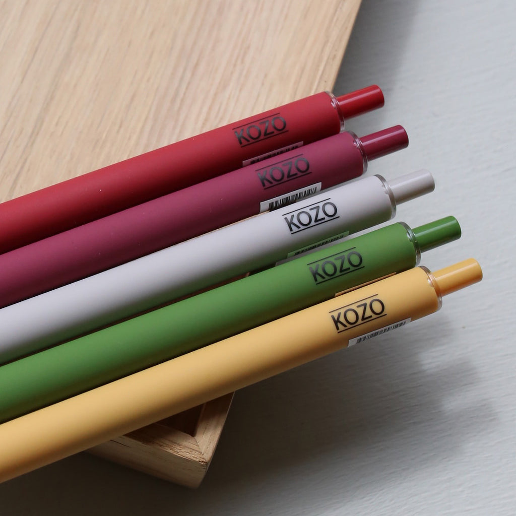 Five KOZO gel pens in berry red, guava red, sand, olive green, and honey, arranged diagonally on a light wood tray over a gray surface.