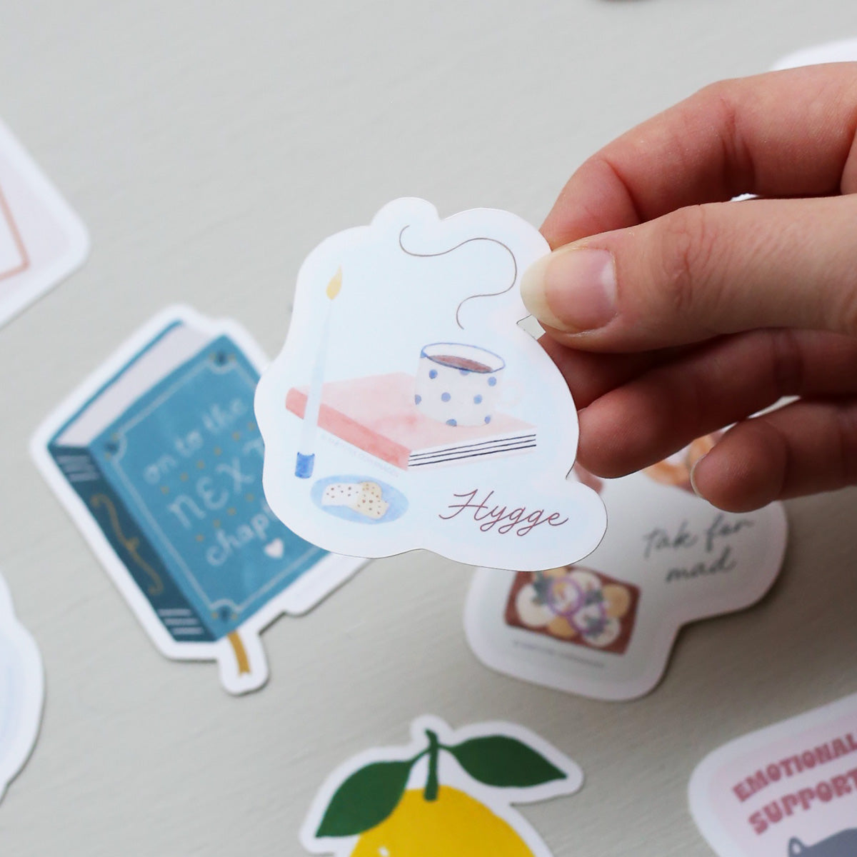 Hand holding a 'Hygge' themed sticker with other decorative stickers on a light surface.