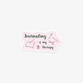 Venyl sticker with 'Journaling is my therapy' text and journal, pen, and jar illustrations on a pink background.