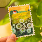 Hand holding a vinyl sticker of a landscape with flowers and a house, surrounded by green leaves.