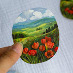 Hand holding a round sticker with a landscape design featuring red flowers and green fields.
